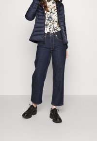 Marineblå quiltet jakke over en blomstret top, parret med brede mørke jeans og kraftige sorte sko. Tekstureret stof og afslappet pasform.