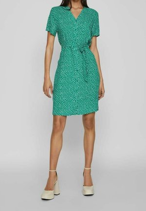 Femme portant une robe verte à manches courtes avec des pois blancs et une ceinture à la taille, associée à des chaussures à talons beiges.