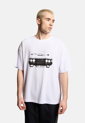 Młody mężczyzna z krótkimi blond włosami ubrany w białą, oversize’ową koszulkę z czarno-białą grafiką zabytkowego samochodu na przodzie.