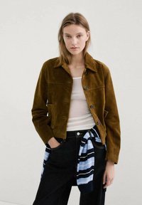 Mujer joven con chaqueta de ante marrón sobre camiseta blanca sin mangas, vaqueros negros y suéter a rayas atado a la cintura, de pie frente a un fondo liso.