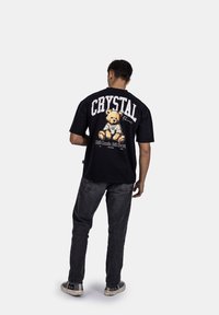 Černé bavlněné tričko s grafikou medvídka a textem "CRYSTAL Paris" na zadní straně. Doplňuje ho tmavé džíny a tenisky.