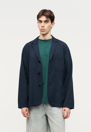 Joven con cabello corto castaño que lleva una chaqueta texturizada azul marino oscura sobre una camisa verde y jeans gris claro, de pie contra un fondo blanco liso.