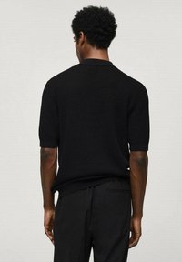 Homme aux cheveux courts et bouclés, portant un polo en maille noire à manches courtes et un pantalon noir, vu de dos sur un fond uni.
