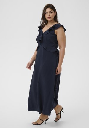 Žena u mornarsko plavoj maxi haljini bez rukava s naborima na izrezu i crnim sandalama s potpeticama i remenčićima.