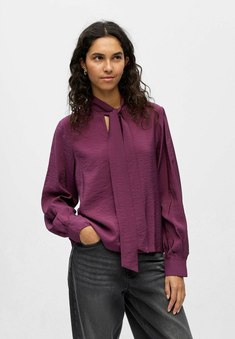 Violette Bluse mit Schlüsselloch-Ausschnitt und Schleifendetail, aus leichtem strukturiertem Stoff, kombiniert mit dunkelgrauen Jeans.