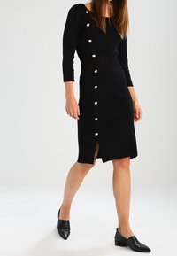 Robe noire à manches longues avec une fente latérale et des boutons dorés sur le côté gauche. Le tissu semble en maille et moulant.