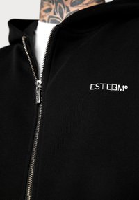 Schwarze Zip-Hoodie aus weichem Stoff, mit einem silbernen Reißverschluss und einem kleinen weißen Logo auf der Brust. Einfaches und modernes Design.