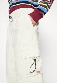 Pantalons cargo beige en tissu léger, avec un cordon de serrage à la taille, des poches latérales et une étiquette de marque visible.