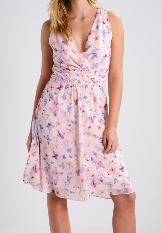 Robe rose pâle sans manches avec imprimé papillons multicolores, encolure en V et taille froncée, portée par une femme debout.