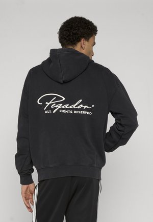 Uomo che indossa una felpa con cappuccio nera con testo "Pegador All Rights Reserved" sul retro, abbinata a pantaloni neri con strisce bianche laterali.