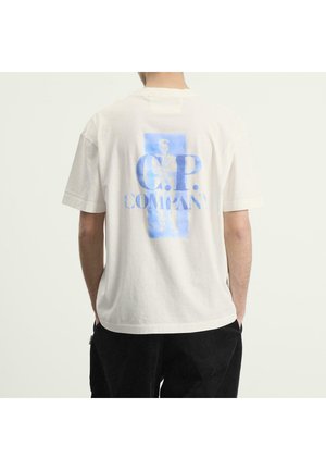 Persona che indossa una t-shirt bianca con la scritta blu "C.P. Company" e una grafica sfocata di una figura sul retro, abbinata a pantaloni neri.