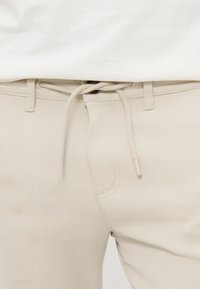 Gros plan d’un pantalon beige avec une taille à cordon et des passants de ceinture, associé à un haut blanc.