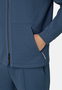 Chaqueta azul con cremallera, hecha de material suave con bolsillos frontales, detalles de cremallera en contraste y un corte holgado. Adecuada para uso casual.