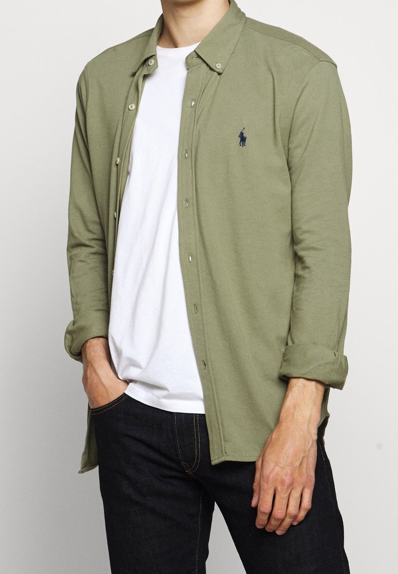 Muž v košili s vyhrnutými rukávy olivové zelené barvy, kterou má přetaženou přes bílé tričko a tmavé džíny, s rukou v kapse, postávající na jednolitém pozadí.