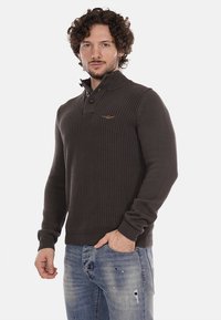 Pullover lavorato a maglia marrone scuro con un piccolo colletto, chiusura a due bottoni e un sottile dettaglio del logo sul petto. Indossato con jeans leggermente strappati di colore azzurro.