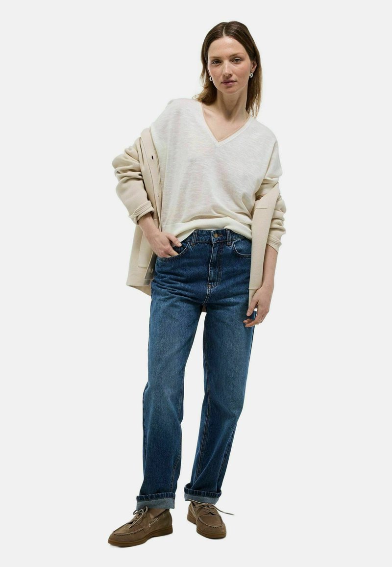 Pull en V beige superposé sur une légère veste beige, associé à un jean denim bleu ample et des chaussures à lacets marron.