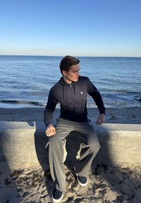 Fiatal férfi tengerészkék kapucnis pulóverben és szürke nadrágban, aki egy beton peremen ül az óceán mellett, tiszta kék ég alatt.