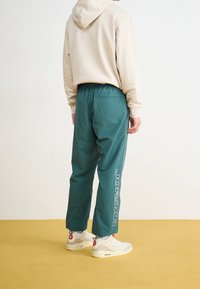 Volné kalhoty tyrkysové barvy s elastickým pasem, s boční kapse a nápisem "JORDAN" v bílé barvě na levé noze.