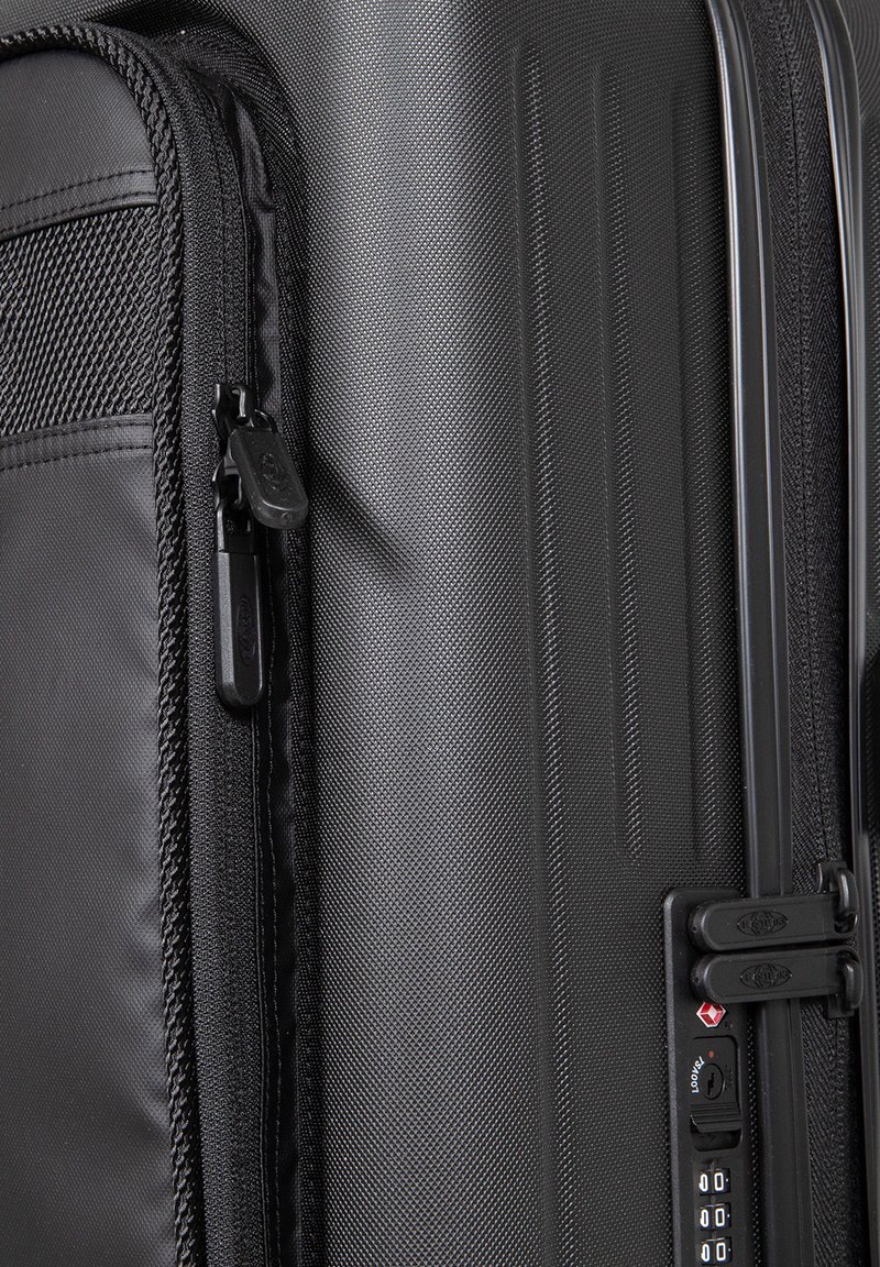Valise rigide noire avec une surface texturée, plusieurs compartiments zippés, un verrou à combinaison intégré et des bords lisses et arrondis.