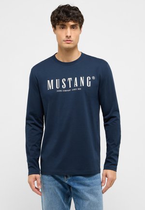 Junger Mann trägt ein navyblaues Langarmshirt mit dem Text "Mustang Jeans Company Since 1932" und hellblaue Jeans vor einem einfarbigen Hintergrund.