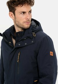 Marineblaue, gefütterte Jacke aus wasserfestem Stoff mit einer Reißverschlusstasche auf der Brust, Kordelzug-Detail und beigem Logo-Patch am Ärmel.