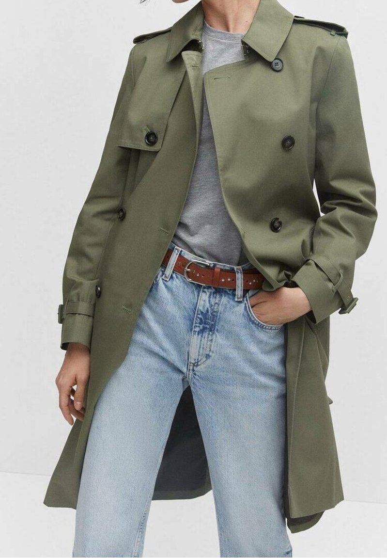Personne portant un trench-coat croisé vert olive par-dessus une chemise grise, rentrée dans un jean bleu clair avec une ceinture marron.