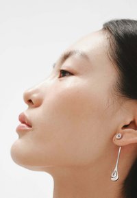 Los pendientes de gota plateados presentan una forma de lágrima con un acabado pulido y un pequeño arete cuadrado con una piedra clara. El diseño es elegante y moderno.