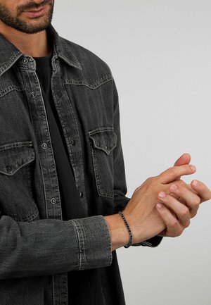 Camicia di denim nera con dettagli dei bottoni, colletto e due tasche sul petto. Un braccialetto di perle con accenti blu è indossato al polso.