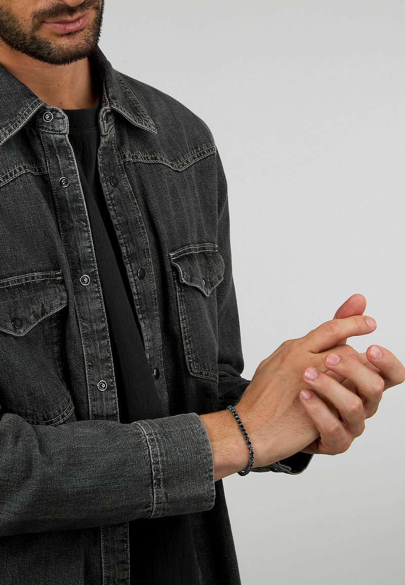Camicia di denim nera con dettagli dei bottoni, colletto e due tasche sul petto. Un braccialetto di perle con accenti blu è indossato al polso.
