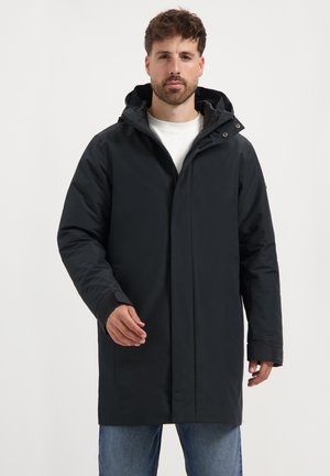 Man in een lange zwarte winterjas met capuchon, over een wit shirt en blauwe jeans, staand tegen een effen witte achtergrond.