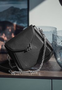 Černá kožená kabelka se strukturovaným designem, s stříbrným řetízkem a kovovým logem. Hladká textura, kompaktní tvar.