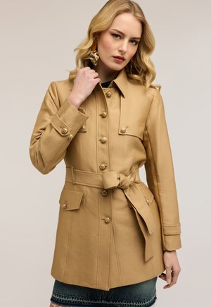 Mujer con un abrigo beige con cinturón, grandes botones y bolsillos con solapa, combinado con una falda vaquera oscura y pendientes llamativos dorados.