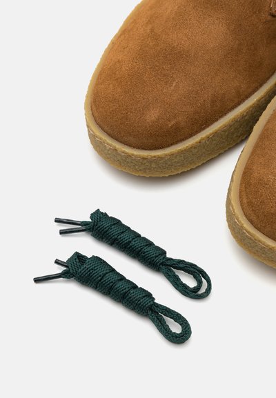 Braune Wildlederschuhe mit runder Spitze und Gummisohle, begleitet von grünen Schnürsenkeln mit gedrehter Textur. Die Schnürsenkel haben schwarze Spitzen.