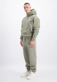 Olijfgroene hoodie met kangoeroezak en logo, gecombineerd met bijpassende joggingbroek. Beide items hebben een relaxte pasvorm en geribbelde boorden. Witte sneakers.