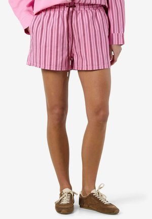 Femeie purtând pantaloni scurți roz cu dungi și șnur, asortați cu teniși maro cu șireturi, stând în picioare pe un fundal simplu.