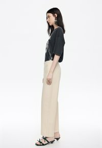 Camiseta gráfica negra de gran tamaño, pantalones rectos beige y sandalias negras con acentos verdes. Diseño simple con ajuste relajado.