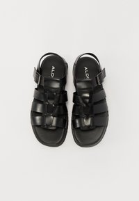 Black sandals with woven straps, a flat sole, and an adjustable buckle on the side. Made of smooth synthetic material with minimal texture.