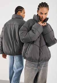 Graue Pufferjacke mit hohem Kragen, aus gestepptem Material und elastischen Bündchen. Auffällige Markenkennzeichnung auf dem Rücken und eine lässige Passform.