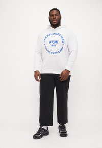 Grauer Kapuzenpullover mit blauem, rundem Logo und Text, kombiniert mit lockeren schwarzen Hosen und schwarzen Sportschuhen mit lila Akzenten.