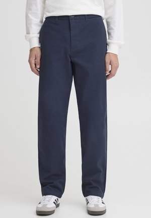 Mand iført marineblå bukser med lige ben, hvid langærmet skjorte og hvide sneakers med sorte striber, stående mod en ensfarvet baggrund.