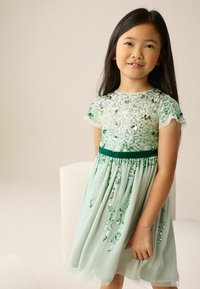 Žalia suknelė su blizgučiais pasipuošusia korsetu ir trumpomis rankovėmis, turinti tekstūruotą, s 흐토누낀음을 흐고 s u r tu s. Juosmuo pabrėžtas audinio dirželiu.