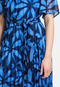 Niebieska sukienka w kwiatowy wzór z plisowaną spódnicą, przewiązana w talii pasującym paskiem. Wykonana z lekkiej, półprzezroczystej tkaniny.