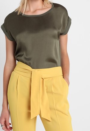 Femme portant un chemisier à manches courtes vert olive et un pantalon jaune taille haute avec une ceinture nouée et les mains dans les poches.