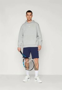 Grå hettegenser, marineblå shorts, hvite joggesko. Holder en tenniskølle med blå accenter. Hud med tatovering synlig på legg.