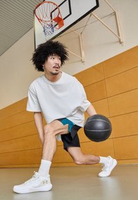 Bílé oversized tričko, černé šortky s tyrkysovými akcenty, černý basketbal a bílé sportovní boty. Pozadí basketbalového hřiště s košem.