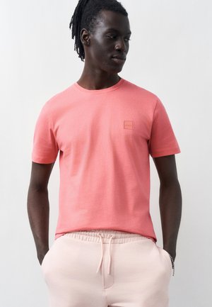 Mand med mørk hud og korte dreadlocks iført en koralrosa kortærmet skjorte og lyserøde snørebukser, hænderne i lommerne.