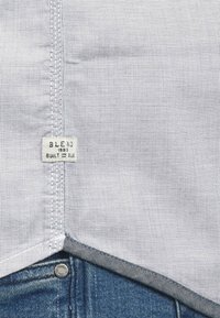 Camicia grigia a motivo con un'etichetta che recita "BLEND 1993 Built in Blue", abbinata a denim blu. Dettagli di cucitura visibili sull'orlo.