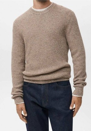 Uomo che indossa un maglione beige a collo tondo in maglia sopra una camicia bianca, abbinato a jeans blu scuro, in piedi davanti a uno sfondo neutro.