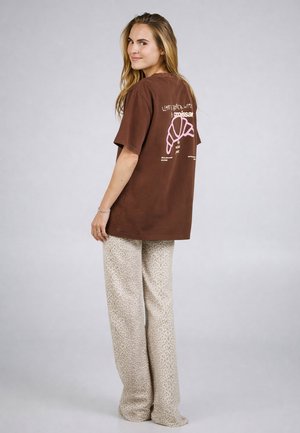 Frau mit langen Haaren, die ein braunes T-Shirt mit rosa Croissant-Grafik und beige, weit geschnittene Hose mit Leopardenmuster trägt, steht seitlich und lächelt.