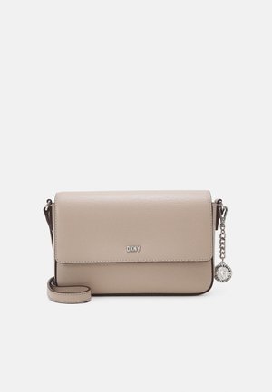 Sac bandoulière beige DKNY avec fermeture à rabat, sangle réglable et breloque logo circulaire argentée attachée à une chaîne latérale.